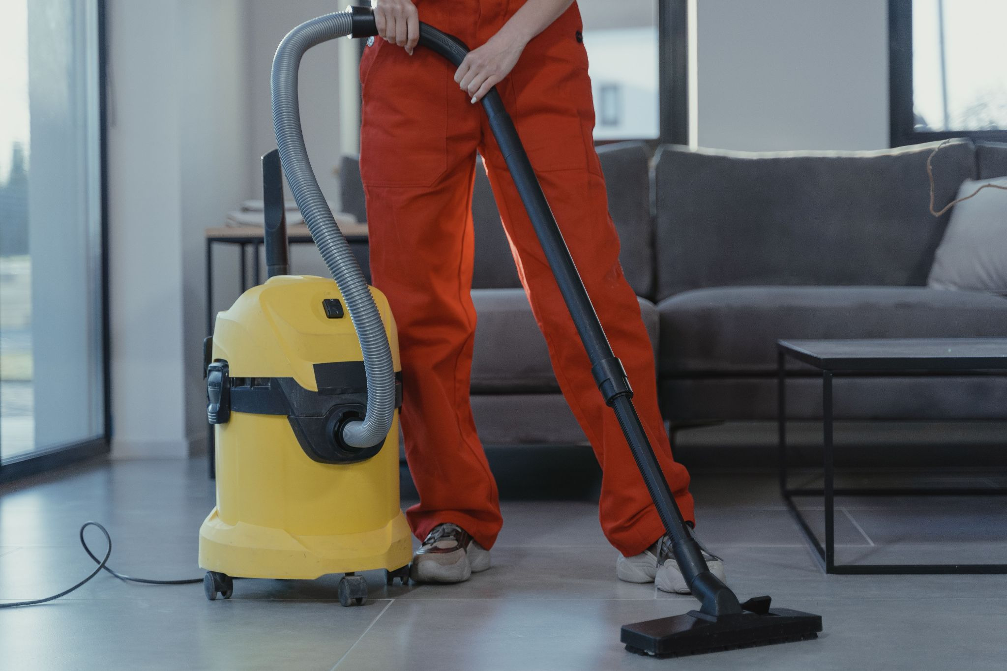 A professional cleaner in red coveralls vacuums a contemporary living room with a yellow vacuum cleaner