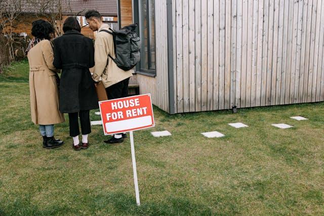 A group of people standing near a “house for rent” sign.